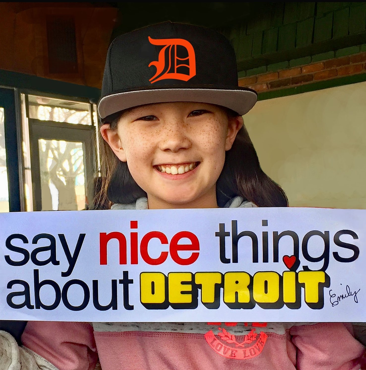 Young Detroit Tigers fan holds up "Say Nice Things About Detroit" bumper sticker designed by Detroit booster Emily Gail.